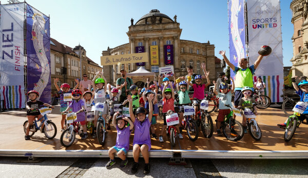  Eine Aufnahme der Teilnehmenden der Laufrad-Tour im Ziel auf dem Theaterplatz. Eine große Gruppe Kinder steht gemeinsam mit einigen Erwachsenen mit Fahrrädern und Fahrradausrüstung auf einem Podest und winken und jubeln in die Kamera. Dahinter wurden Banner mit dem „Sports United“-Logo und dem Schriftzug „Ziel“ aufgebaut. Es gibt auch ein Schild, auf dem „Laufrad-Tour“ steht. Im Bildhintergrund sind in der Mitte das Opernhaus, rechts ein Teil der Petrikirche und links das Gebäude der Kunstsammlungen Chemnitz zu erkennen. Der Himmel ist blau und wolkenlos.