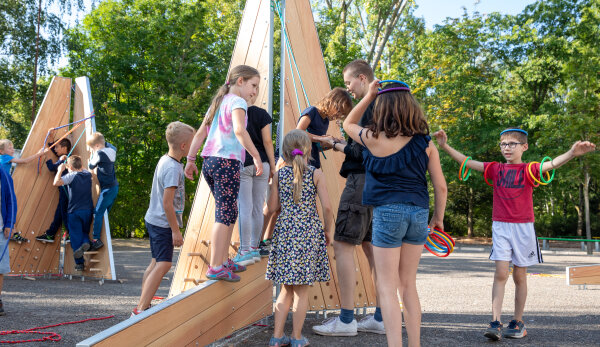 Eine Aufnahme des Schulhofs der Albert-Einstein-Grundschule. Im Vordergrund ist eines der Objekte, die zum Gesamtobjekt „Playwall“ gehören, abgebildet. Das Objekt besteht aus trapezförmigen Platten aus hellem Material, die stehend und liegend angeordnet sind. Viele Schülerinnen und Schüler stehen und klettern auf dem Objekt und stehen daneben. Links am Bildrand ist ein zweites ähnliches Objekt abgebildet; auch dieses wird von den Kindern kletternd erkundet. Rechts steht ein Junge, der die Arme ausbreitet und auf dem Kopf und um die Arme herum bunte kleine Reifen balanciert. Im Hintergrund befinden sich Bäume.