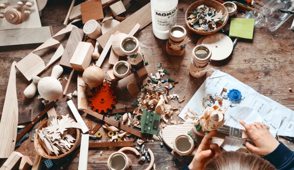 Eine Nahaufnahme einer Tischfläche aus Holz. Darauf liegen durcheinander verschiedene Bastelmaterialien, Holzstücke, Kleinteile und bunte Zahnräder. Außerdem sind ein Bogen Papier mit Notizen und Zeichnungen sowie zwei Kinderhände in der unteren rechten Ecke erkennbar.